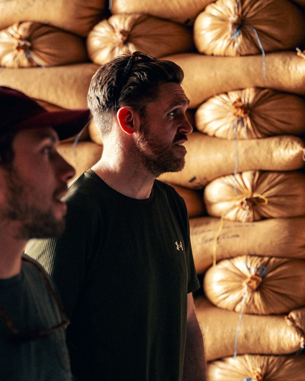 Two men standing in front of a wall of stacked burlap bags.